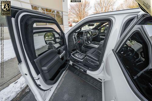 Interior view of an open Mastiff, showing the driver's side and rear door open with visible seats, dashboard, and controls. Outside, there's a building, window reflection, and a curb with snow.