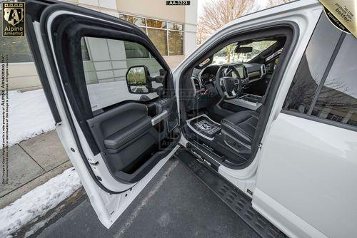 Driver side view of a white Mastiff with both driver and passenger side doors open, revealing the interior. Snow is visible on the ground outside, and a building with glass windows is in the background.