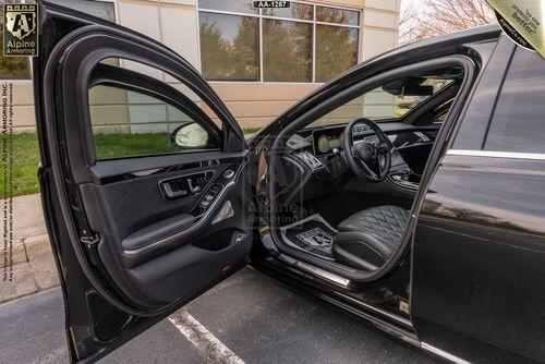 Black luxury Mercedes-Benz S580 Executive sedan with the driver's door open, revealing a quilted leather interior and modern dashboard. The car is parked outside a building with large bulletproof windows.