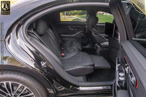 The interior of an armored black Mercedes-Benz S580 Executive sedan shows leather seats, a screen mounted behind the passenger seat, and a clear view of the exterior through the open door. Various branding elements are visible on the image.