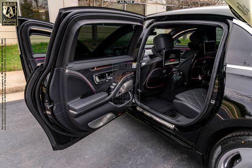 A black armored Mercedes Maybach with open rear passenger doors showcasing a premium interior with leather seats, mood lighting, and entertainment screens on the back of the front seats. The car is parked outdoors.