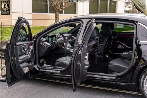 A Mercedes-Benz Maybach with both front and rear side doors open, revealing a sleek, modern interior with leather seats and a high-tech dashboard. The car is parked outside a building with large windows.