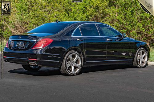 A black luxury armored black Mercedes S550 sedan is parked on a pavement with a background of green foliage.