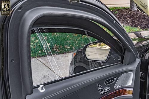 Close-up view of a Mercedes-Benz S550's interior door panel and side mirror, with the bulletproof glass window slightly open. The exterior shows concrete pavement, grass, and the front portion of another vehicle.