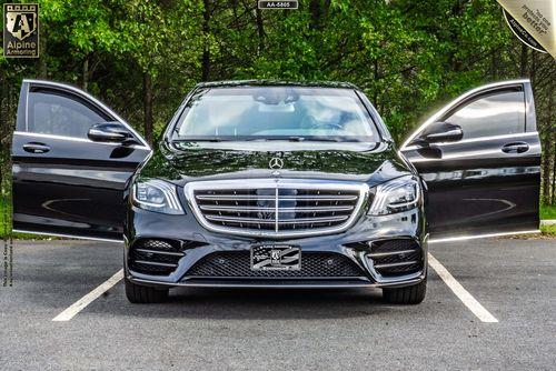 A black luxury Mercedes-Benz 560 with both front doors open is parked in an outdoor lot, framed by a green, wooded background.