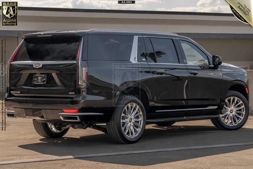 A black, large luxury armored Cadillac Escalade ESV Premium Luxury SUV with tinted windows parked on a concrete surface under a blue sky with clouds. The vehicle has chrome accents and polished alloy wheels.