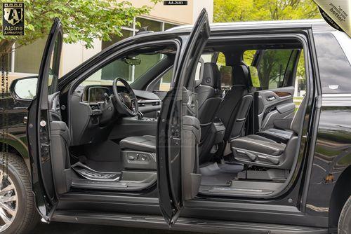 A black armored Cadillac Escalade ESV Premium Luxury SUV with all doors open, showcasing three rows of gray leather seats and a modern dashboard, parked outdoors with some greenery visible in the background.