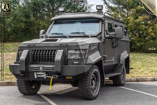 An armored SWAT APC CUDA with rugged tires and a gray exterior is parked on a paved area beside grassy surroundings. The vehicle has a winch attached to the front bumper.