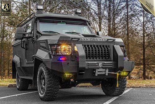 A black armored SWAT APC CUDA with rugged tires and a robust exterior is parked on a lot with trees in the background.