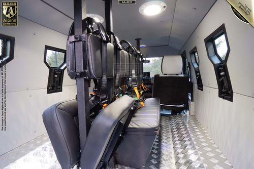 Interior of an armored SWAT APC CUDA, with black padded seats along the left side, metallic flooring, and a driver's seat at the front. The ceiling is lined with light fixtures and the walls have small windows.