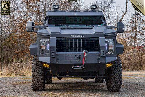 Front view of a black armored vehicle with rugged tires and robust protective features, labeled "Pit-Bull VX." The vehicle is parked outdoors in a wooded area.