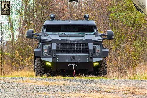 Front view of a black armored Pit-Bull VX with rugged tires parked on a gravel path, surrounded by tall brown grass and leafless trees. The vehicle has protective bumpers and roof-mounted lights.