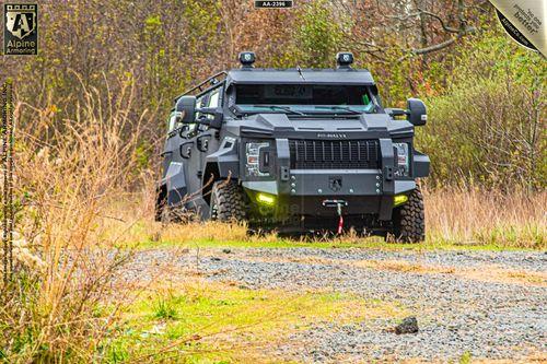 An armored Pit-Bull VX is seen driving on a rocky and grassy terrain, surrounded by dry vegetation. The vehicle is large, heavily fortified, and painted in a dark color.