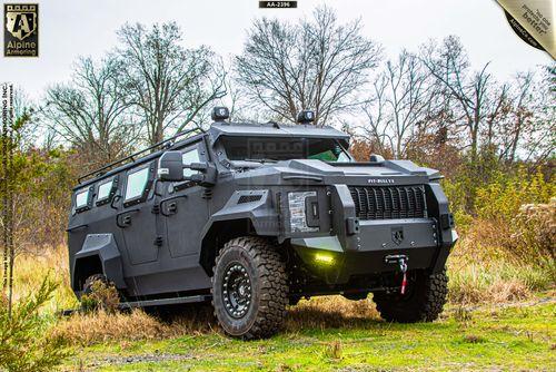 An armored Pit-Bull VX is parked on grass with trees in the background. The vehicle has a rugged design with large tires and is equipped with lights and winch.