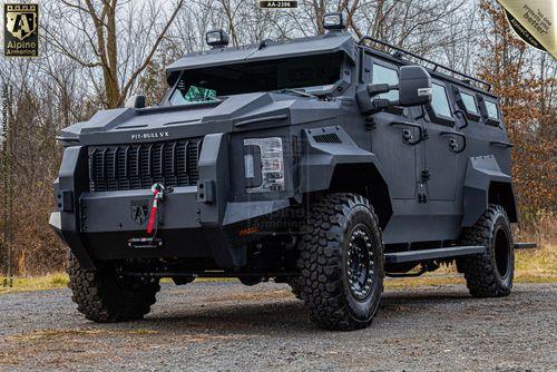 A black, heavily armored Pit-Bull VX with rugged tires and a prominent front grille is parked outdoors, surrounded by trees.