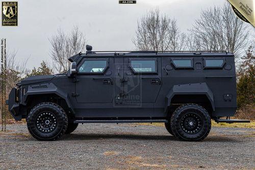 Side view of a black Alpine Armoring Inc. armored Pit-Bull VX parked on a gravel surface with leafless trees in the background.