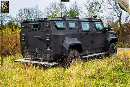 A black armored Pit-Bull VX  is parked in a grassy area. It is equipped with rugged tires, reinforced windows, and several antennae. Trees and a cloudy sky are in the background.