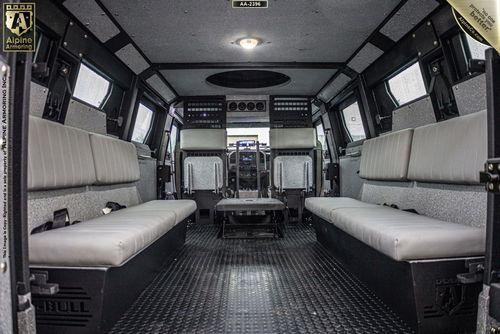 Interior of an armored Pit-Bull VX with two rows of gray bench seats along the sides, gray walls, and a control panel at the front.