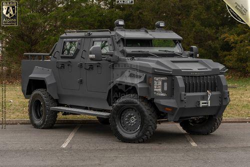 A black, heavily armored black Pit-Bull VXT vehicle parked on a paved area in front of trees. The vehicle features rugged tires, protective plating, and roof-mounted equipment.