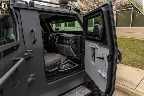 An armored black Pit-Bull VXT with an open driver's side door, showcasing the interior seating and dashboard. The exterior appears matte black, and a building is visible in the background.