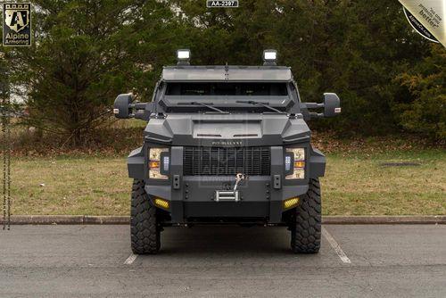 A front view of an Alpine Armoring Pit-Bull VXT armored vehicle parked in an outdoor setting with trees in the background.