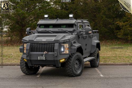 A rugged, black armored black Pit-Bull VXT is parked on a paved surface with a background of trees. It has large tires, multiple lights, and robust protective plating.