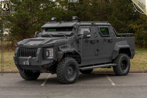 A matte-black armored black Pit-Bull VXT with large tires is parked on asphalt, facing slightly to the left. Trees are visible in the background.