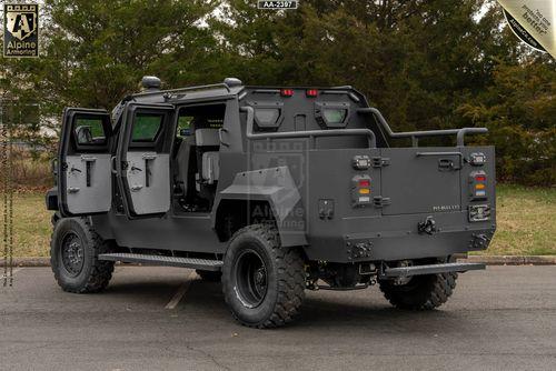 A black, armored Pit-Bull VXT is parked outdoors. The rear and side doors are open, revealing a detailed interior. The vehicle is surrounded by greenery and pavement.