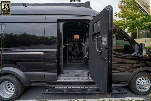 The passenger side view of a black armored Pointer van with an open thick steel door displaying the Alpine Armoring logo. The interior is minimally visible through the open door. The van is in a parking lot with trees in the background.