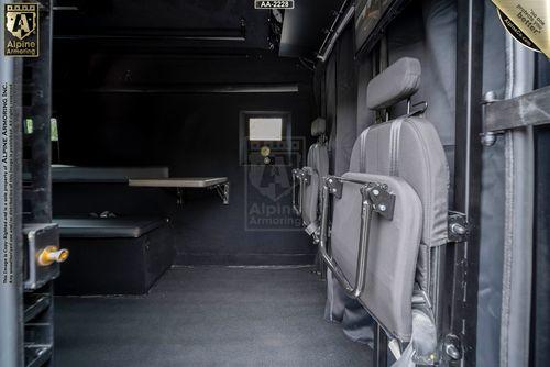 Interior of an armored Ford Transit Pointer Van with seating and a small work desk along the left wall. The seats have metal frames and are attached to the right wall. The environment is enclosed and utilitarian.