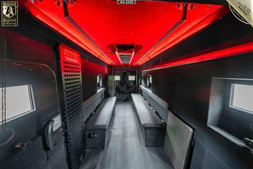 Interior of a dark, armored Pointer Van with red ceiling lights, two parallel benches, small rectangular windows, and secure compartments. The walls and floor feature utilitarian design elements.