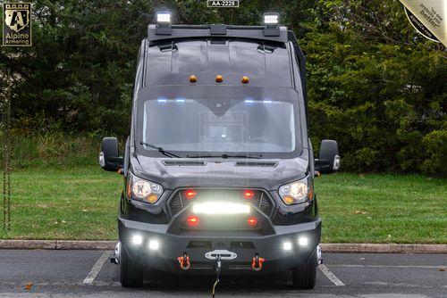 A black Ford Transit Pointer Van equipped with front-facing lights, a winch, and a robust grille guard is parked on a paved surface with green foliage in the background.