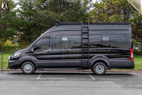 Driver side view of a black SWAT VAN - Pointer  parked in a parking lot. The van features tinted windows, a side ladder, and roof modifications. Trees and grass are visible in the background.