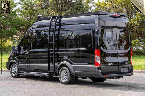 A black, large SWAT VAN - Pointer Based on Ford Transit is parked in an outdoor setting with trees in the background. The van features a rooftop rack, side ladder, and various modifications for potential off-road or adventure use.