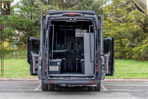 A Ford Transit Pointer Van with its rear doors open, revealing an armored interior with a workstation setup, including a chair, desk, and equipment. The exterior shows 'Alpine Armoring' branding.