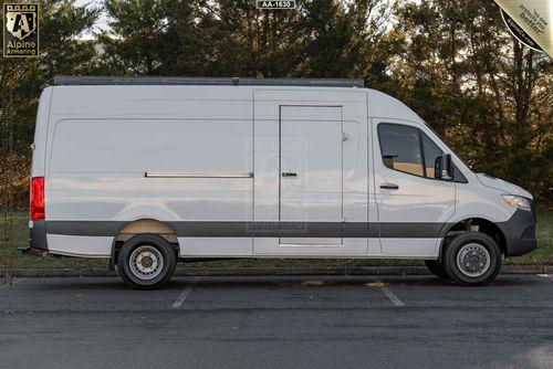 A white Mercedes-Benz Sprinter van with a sliding side door is parked in a parking lot, next to a row of trees.