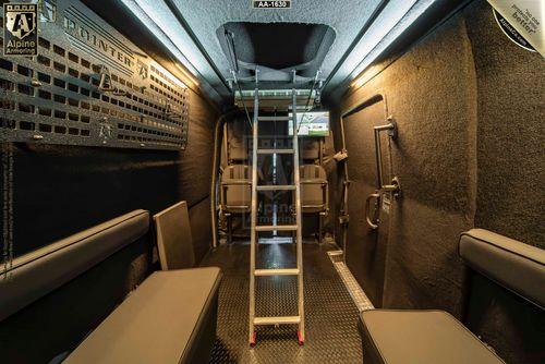 Interior view of a mobile tactical vehicle, the SWAT Van - Pointer, featuring secured seating, a central retractable ladder, padded walls, bright overhead lighting, and various secured equipment. Alpine Armoring branding visible.