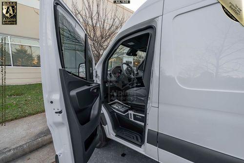 An open driver's side door of a white SWAT Van - Pointer displaying the steering wheel, dashboard, and front interior seats. The van is parked near a beige building with trees and grass in the background.