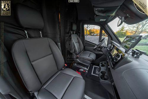 Interior of an armored SWAT Van - Pointer showing two front seats, a dashboard with a mounted device, and cup holders, with a logo on the seat and ceiling. Trees are visible through the windshield.