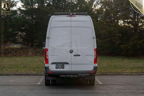 A rear view of a parked white SWAT Van - Pointer® Based on Mercedes-Benz Sprinter  with closed back doors, set against a backdrop of trees and grass. The van has a visible license plate and Alpine Armoring branding.