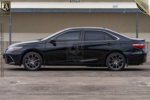 Driver side profile of a black bulletproof Toyota Camry parked in an outdoor setting, seen from the side. The vehicle has tinted windows and aftermarket wheels.