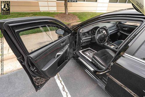 A black armored Toyota Camry with its front passenger door open, showing the interior with black seats, a steering wheel, and dashboard. The car is parked in front of a beige building.