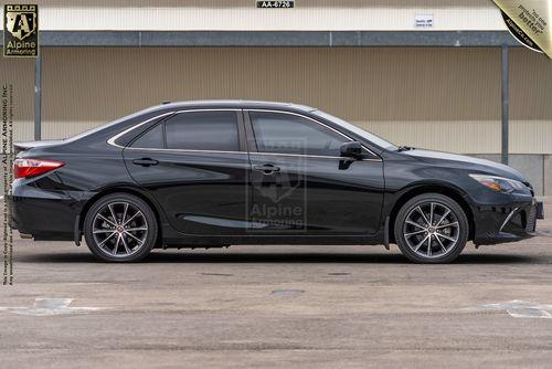 Side view of a black, four-door arrmored Toyota Camry with tinted windows and alloy wheels, parked on a concrete surface.