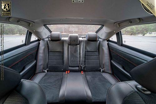 Interior view of a bulletproof Toyota Camry showcasing the rear passenger seats with black leather upholstery and red stitching. The back window and partial rearview of the exterior are visible through the windows.