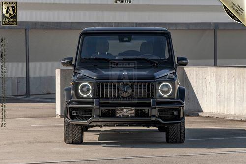 A black armored Mercedes-Benz G63 AMG SUV is parked in an outdoor urban area. The front view displays the grille, headlights, and license plate.