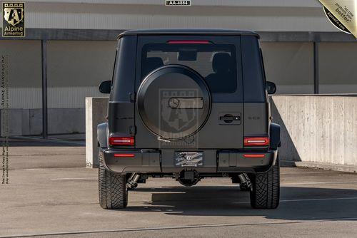 Rear view of a matte black  Mercedes G-Class SUV parked in an urban setting, showcasing its spare tire cover and tail lights.