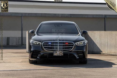 Front view of a black Mercedes-Benz S580 4MATIC from Alpine Armoring, featuring integrated red and blue emergency lights in the grille. The vehicle is parked in an outdoor setting with a concrete wall and building in the background.