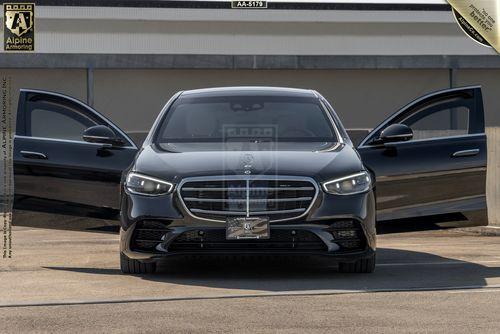 A front view of a black Mercedes-Benz S-Class (S580 4MATIC) from Alpine Armoring with its doors open, showcasing the luxury sedan's design and spacious interior. The vehicle features a distinctive grille and sleek headlights.