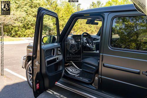 An open driver's door of a black G63, revealing a black interior with visible branding from Alpine Armoring. The vehicle is parked outdoors near trees.