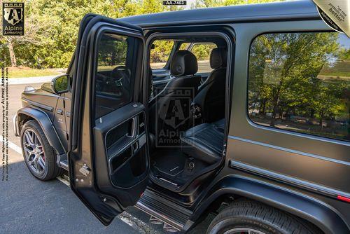 A black armored Mercedes-Benz G63 SUV with its rear passenger door open, revealing black leather seats and a forested background. The vehicle appears modified for armored functionality by Alpine Armoring.